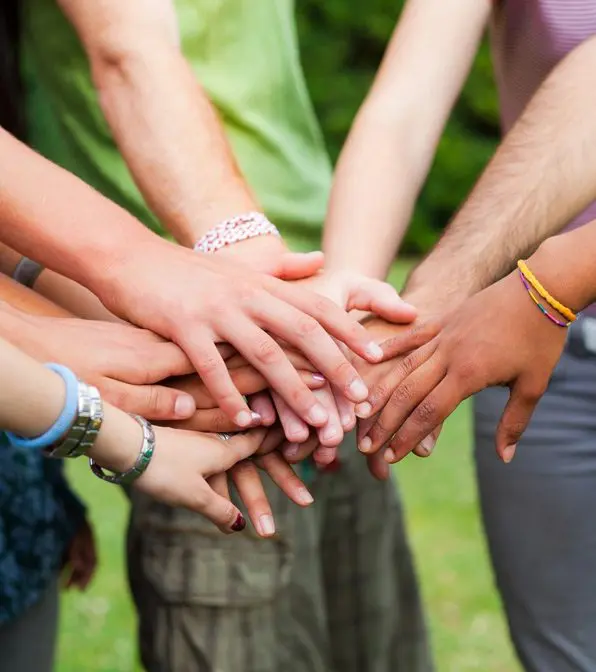 A group of people stand in a circle with their hands stacked together, signifying unity and teamwork. The background is blurred greenery.