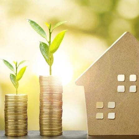 Two stacks of gold coins, each topped with a small plant, sit next to a cardboard house model, symbolizing financial growth and real estate investment.