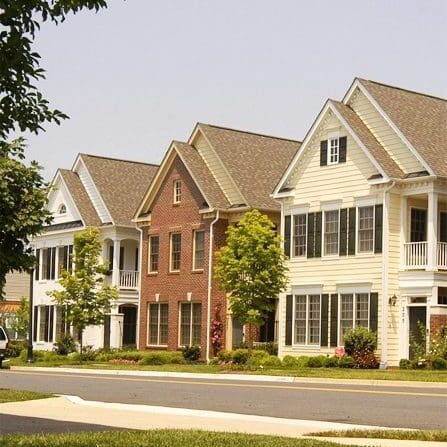 A row of three suburban houses, one brick and two with siding, along a residential street with greenery and well-maintained lawns.