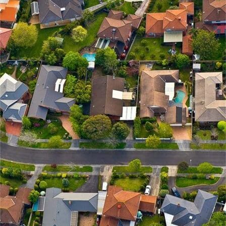 Aerial view of a suburban neighborhood with houses, green lawns, trees, and streets. The houses vary in size and design, and some have swimming pools in their yards.