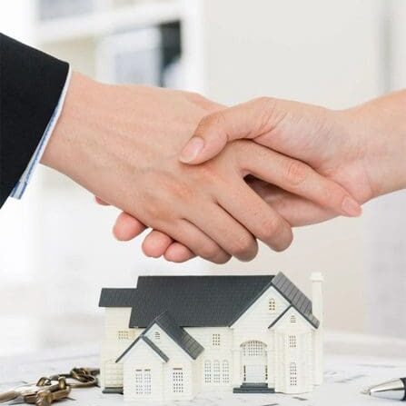 Two people shaking hands over a table with a small model of a house and keys.